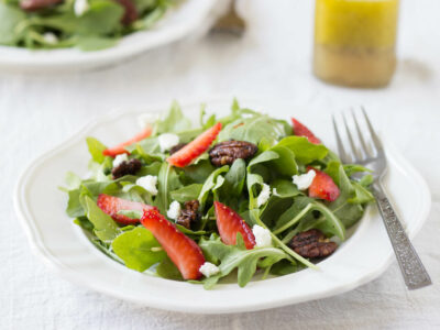 strawberry arugula salad with candied pecans and goat cheese on white plate with dressing