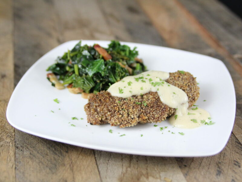 Wide shot of pecan encrusted chicken breast with cauliflower gravy on a white square-like plate on a wood table