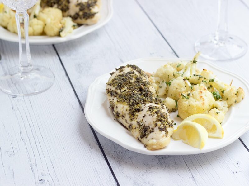 Chicken breast with Greek spices with a side of cauliflower and lemon slices on a white plate and white wood background - wide shot