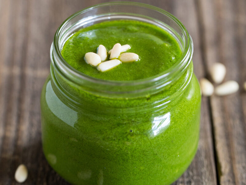 Clear glass jar of Lemon-Basil Pesto with sprinkles of pine nuts on a wood table - wide shot.