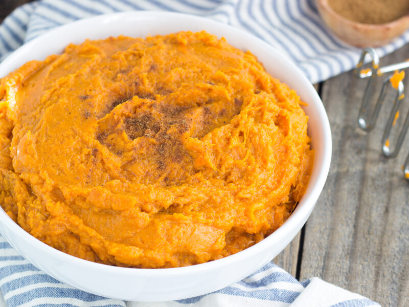 Wide shot of mashed sweet potatoes in a white bowl on a gray weed background.