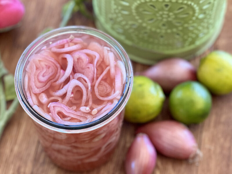 Wide shot of a large jar of pickled shallots with whole limes and shallots visible next to the jar.