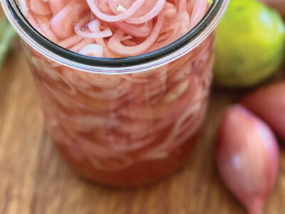 Vertical image of a large jar of pickled shallots with a fork full on the table