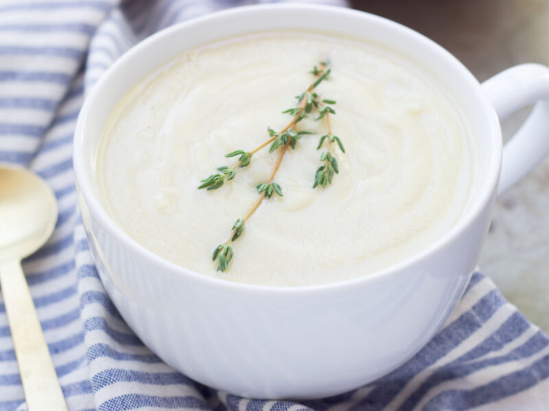 Wide shot of celery root soup in a large white mug topped with a sprig of fresh thyme set near a blue and white napkin and a silver soup spoon