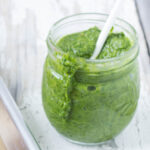 Wide shot of arugula pesto in a glass jar with a white spoon