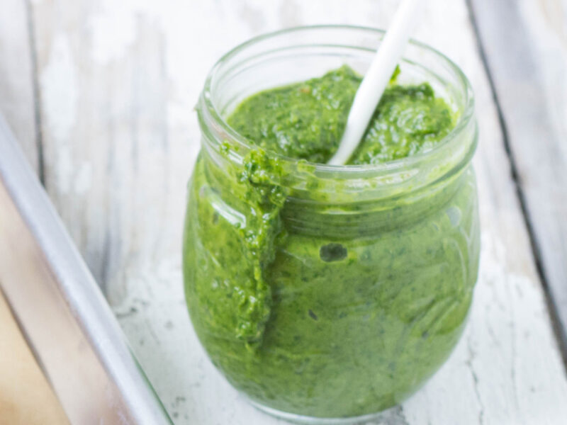 Wide shot of arugula pesto in a glass jar with a white spoon