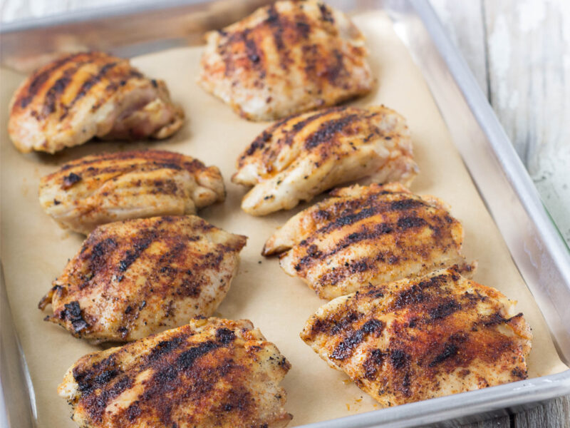 Wide image of eight barbecued chicken thighs with grill marks on a sheet pan with parchment paper.