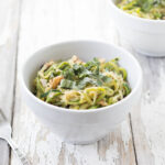 Horizontal image of Bacon-Basil Zucchini "Pasta" in a white bowl with a fork to the side on a white distressed wood background.