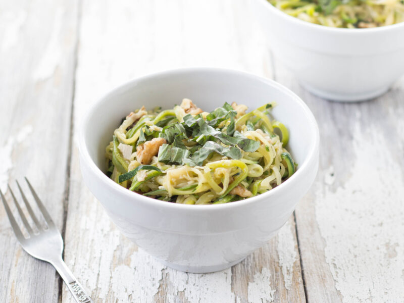 Horizontal image of Bacon-Basil Zucchini "Pasta" in a white bowl with a fork to the side on a white distressed wood background.