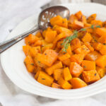 Roasted butternut squash in an oval bowl garnished with a sprig of fresh thyme and an antique silver serving spoon.