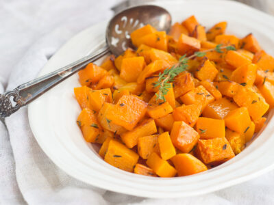 Roasted butternut squash in an oval bowl garnished with a sprig of fresh thyme and an antique silver serving spoon.