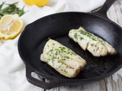 Cooked tarragon whitefish in seasoned cast iron pan on a light wood background.