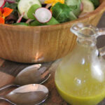 Homemade Italian Dressing in a classic clear glass cruet with a glimpse of colorful salad in a wood bowl in the background.