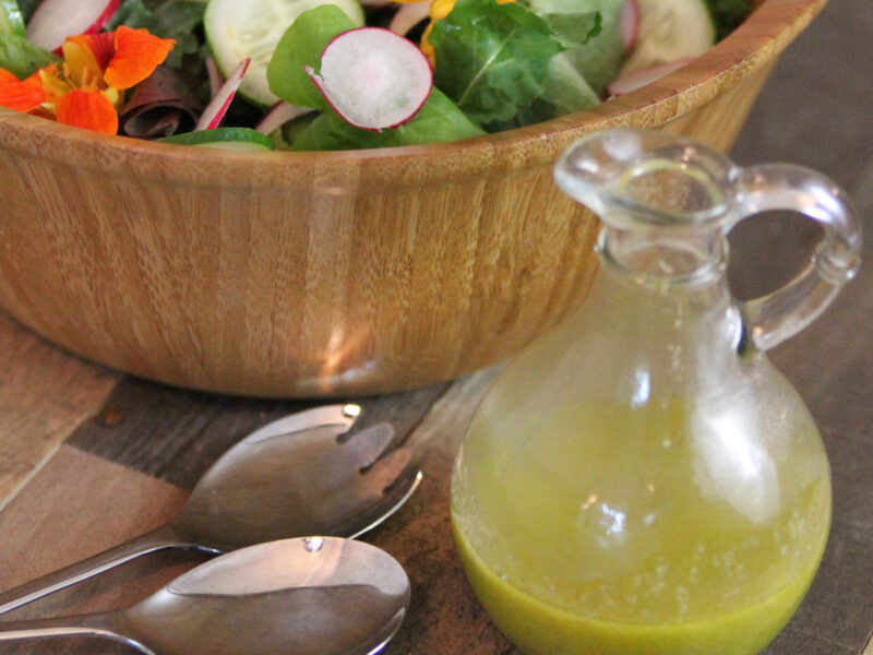 Homemade Italian Dressing in a classic clear glass cruet with a glimpse of colorful salad in a wood bowl in the background.