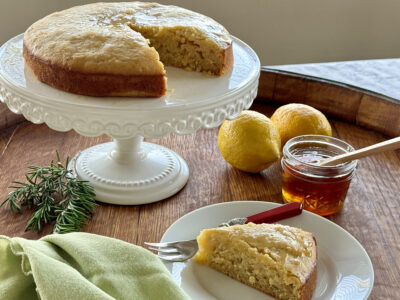 Lemon-Rosemary Olive Oil Cake on white cake stand with slice removed on small white plate (lemons, honey, and rosemary in the background)