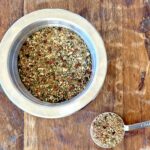 Dried herb steak seasoning mixture in a small stainless steel prep bowl with one tablespoon full measured out on a wood background.