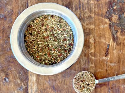 Dried herb steak seasoning mixture in a small stainless steel prep bowl with one tablespoon full measured out on a wood background.