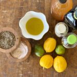 Overhead view of ingredients like lemons, limes, vinegars, oil, salt and paper to make a basic vinaigrette with finished vinaigrette in a small fluted white bowl on a wood background.