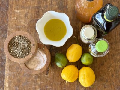 Overhead view of ingredients like lemons, limes, vinegars, oil, salt and paper to make a basic vinaigrette with finished vinaigrette in a small fluted white bowl on a wood background.