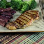 Image of grilled New York strip steak with grill marks on a plate with purple sweet potato fries and broccoli.