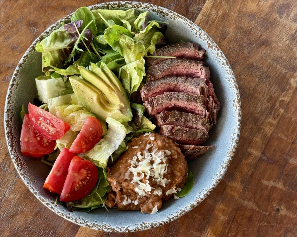 Perfectly medium-rare sliced flank steak with refried beans and a mixed green salad with tomatoes and avocodo in a distressed gray bowl on a wood background.