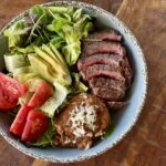Perfectly medium-rare sliced flank steak with refried beans and a mixed green salad with tomatoes and avocodo in a distressed gray bowl on a wood background.