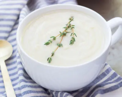 Wide shot of celery root soup in a large white mug topped with a sprig of fresh thyme set near a blue and white napkin and a silver soup spoon
