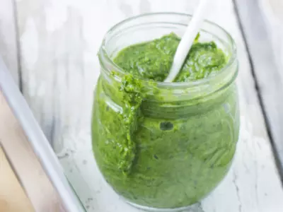 Wide shot of arugula pesto in a glass jar with a white spoon