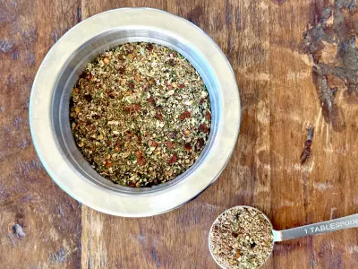 Dried herb steak seasoning mixture in a small stainless steel prep bowl with one tablespoon full measured out on a wood background.