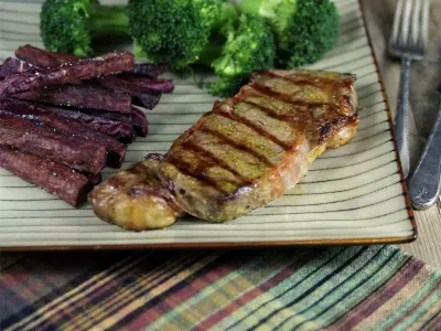 Image of grilled New York strip steak with grill marks on a plate with purple sweet potato fries and broccoli.