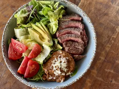 Perfectly medium-rare sliced flank steak with refried beans and a mixed green salad with tomatoes and avocodo in a distressed gray bowl on a wood background.